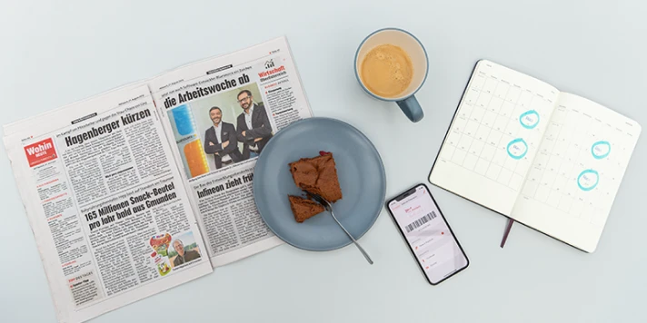 Aufgeschlagene Zeitung, Kalender, Smartphone und Kaffee und Kuchen.