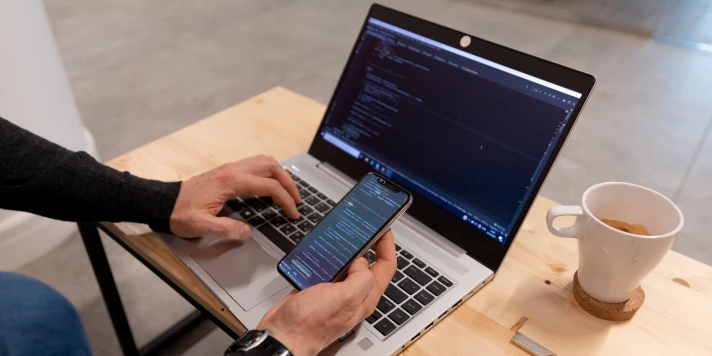 Person bedient mit linker Hand Tastatur eines Laptops und hält Smartphone in rechter Hand.