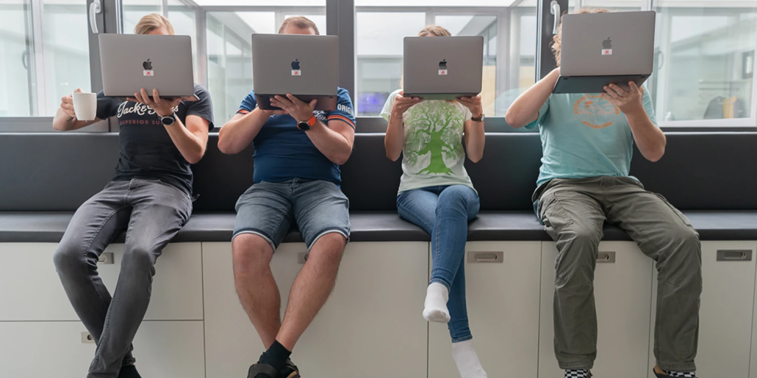 Vier Mitarbeiter*innen sitzen nebeneinander auf Gang und halten Laptop vor dem Gesicht.