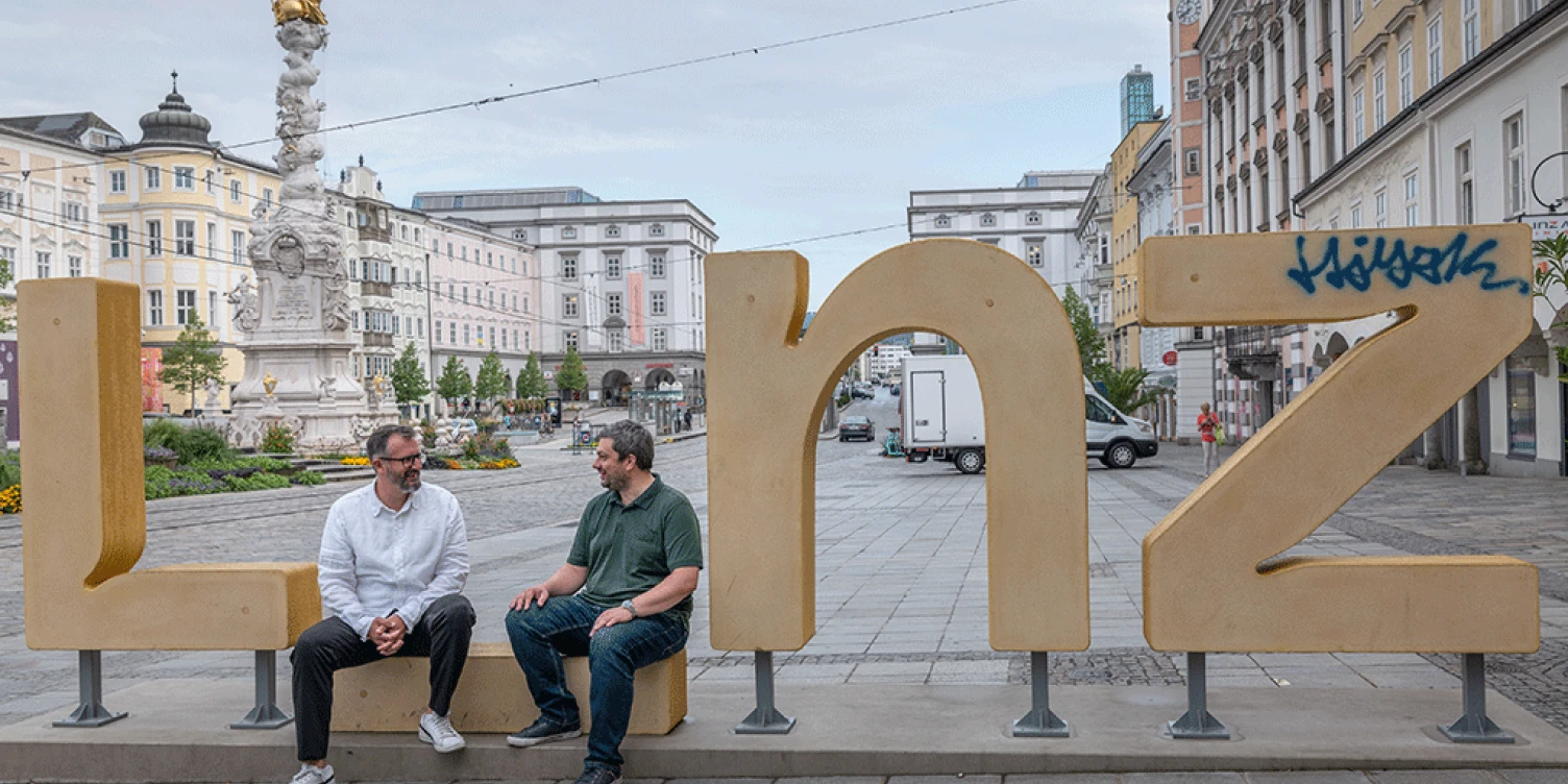 CEO Roland Sprengseis und COO Christian Neudorfer sitzen auf Linz Logo am Linzer Hauptplatz.