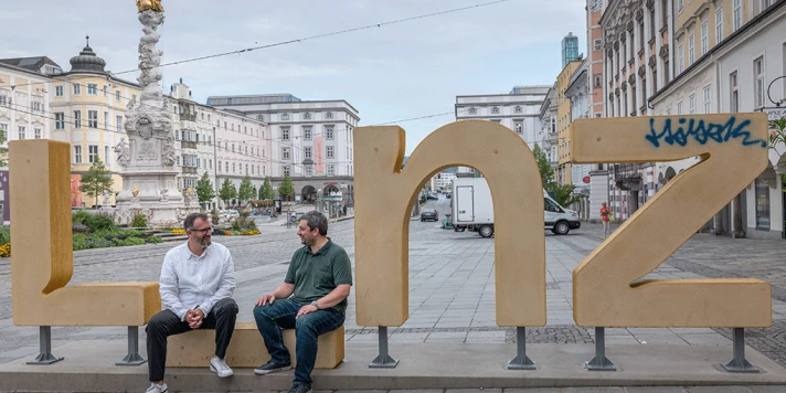 CEO Roland Sprengseis und COO Christian Neudorfer sitzen auf Linz Logo am Linzer Hauptplatz.