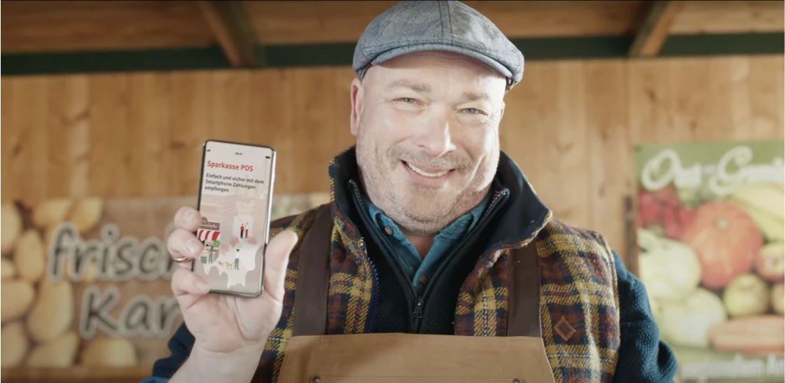 Market stall operator smilingly shows smartphone with Sparkasse POS app.