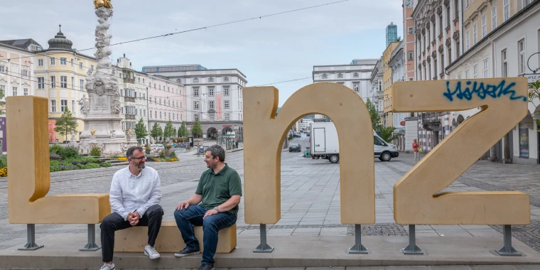 Roland Sprengseis und Christian Neudorfer sitzen am Linz-Logo am Linzer Hauptplatz.