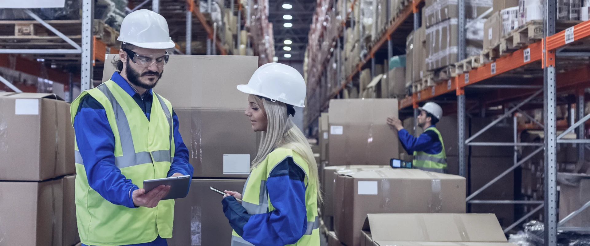 Man and woman in work clothes stand in a high-bay warehouse and look at a tablet.