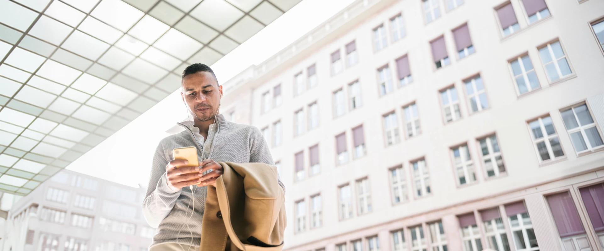 Ein junger Mann steht vor einem hellen Stadtgebäude und blickt auf sein Smartphone. Er trägt Kopfhörer, einen grauen Pullover und hält einen beigen Mantel über dem Arm.