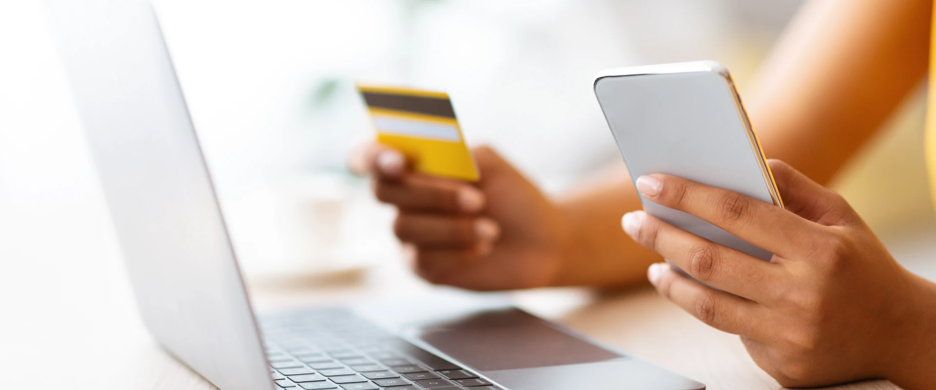 Nahaufnahme einer Person, die vor einem Laptop sitzt, eine gelbe Kreditkarte in der einen und ein Smartphone in der anderen Hand hält – symbolisch für Online-Zahlung oder Mobile Banking.