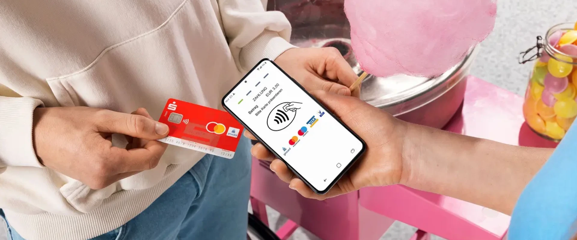 Eine Person hält eine rote Sparkassen-Debitkarte an ein Smartphone, das als mobiles Bezahlterminal fungiert. Im Hintergrund ist eine rosa Zuckerwattemaschine zu sehen.