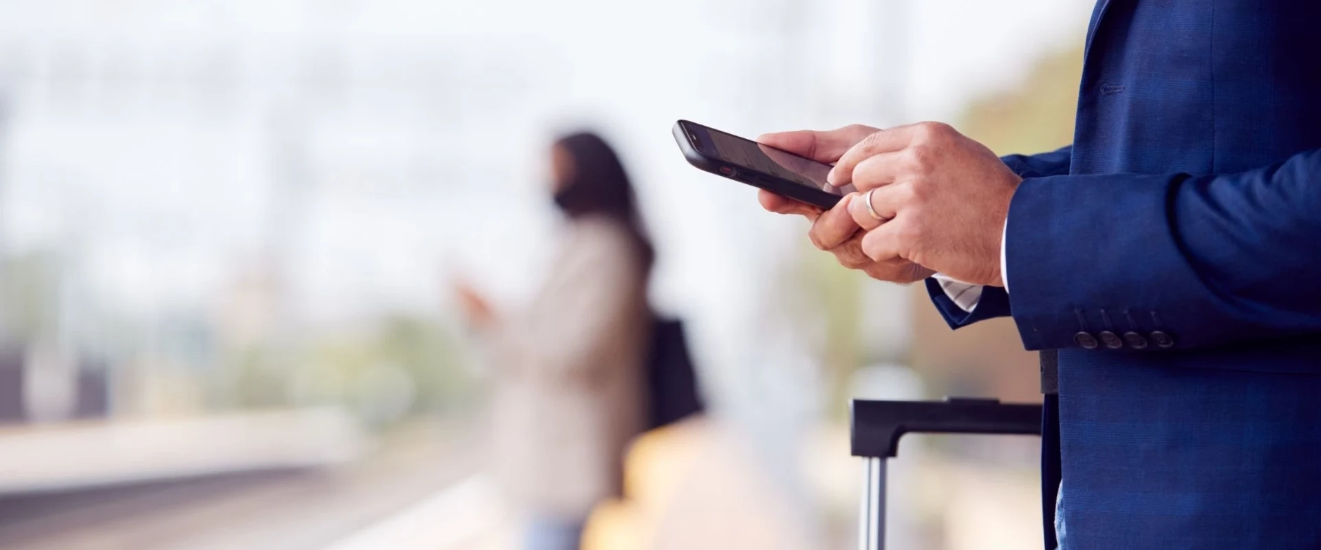 Seitenansicht eines Mannes im blauen Sakko, der am Bahnsteig auf sein Smartphone tippt. Im Hintergrund ist verschwommen eine weitere Person und ein Rollkoffer zu sehen.